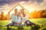 Kleinfamilie sitzt auf einer Wiese und hält sich ein Dach über den Kopf.